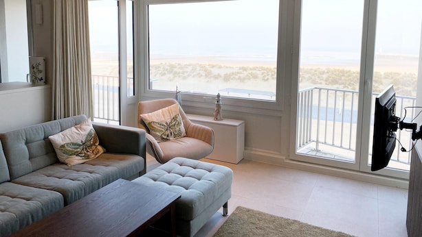 A modern living room with a grey sofa, a small table, and a view of the beach through large windows. - Apartment in Nieuwpoort Near the Beach (Nieuwpoort)