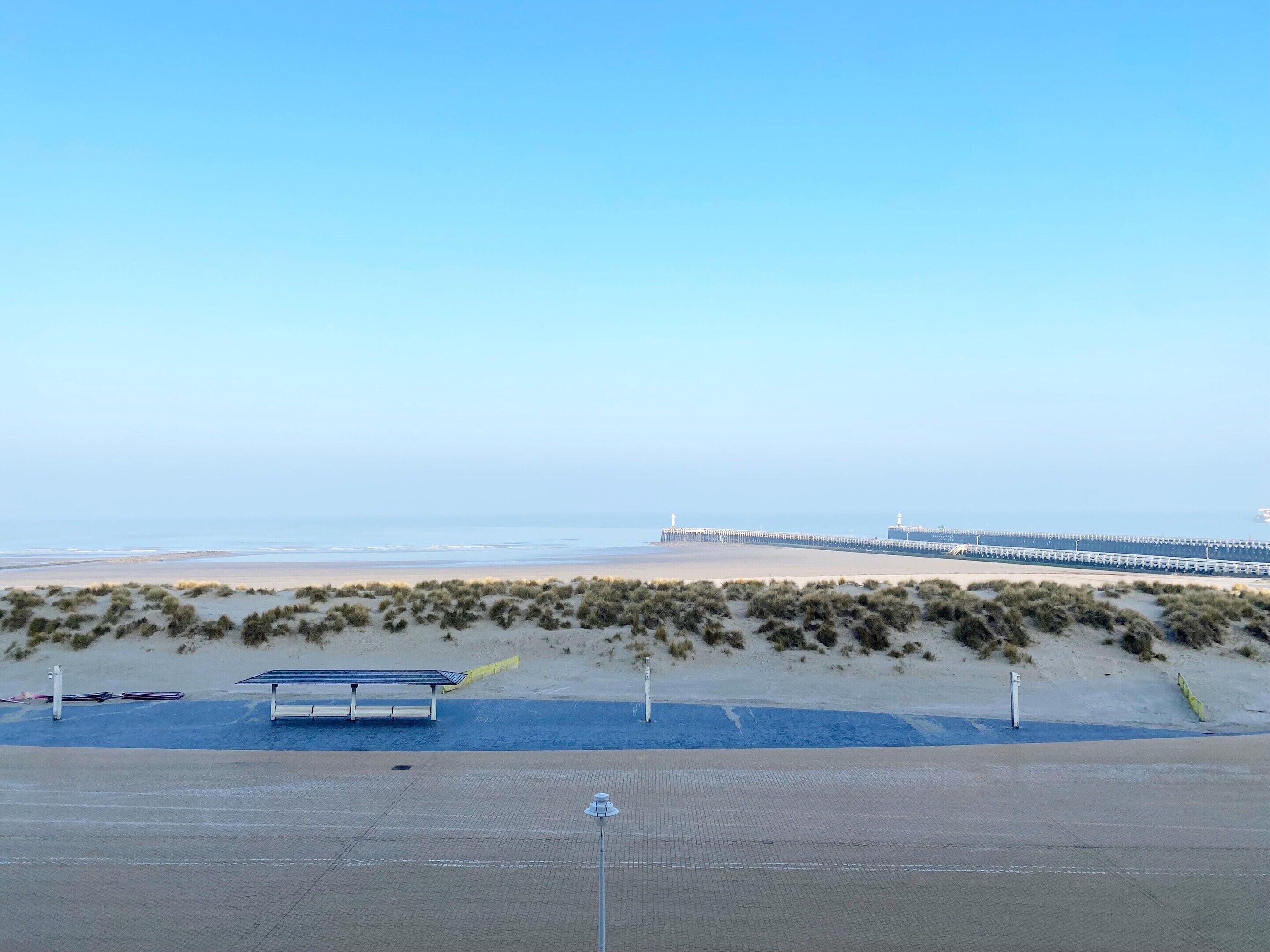 Une plage avec une longue jetée qui s’avance dans la mer, une zone sablonneuse à la végétation clairsemée et un espace de repos couvert équipé d’un panneau solaire.