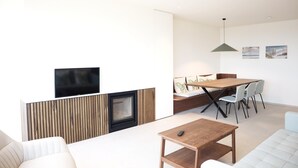 A modern living room with a sofa, a wooden coffee table, a dining table with chairs, a fireplace, and a flat-screen TV mounted on the wall. - Apartment in Nieuwpoort Near the Beach (Nieuwpoort)