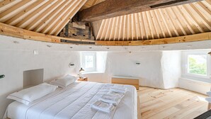 Ein Schlafzimmer mit schrägen Wänden, Holzbalken, einem Bett mit weißer Bettwäsche, einem Nachttisch und einem Fenster.
