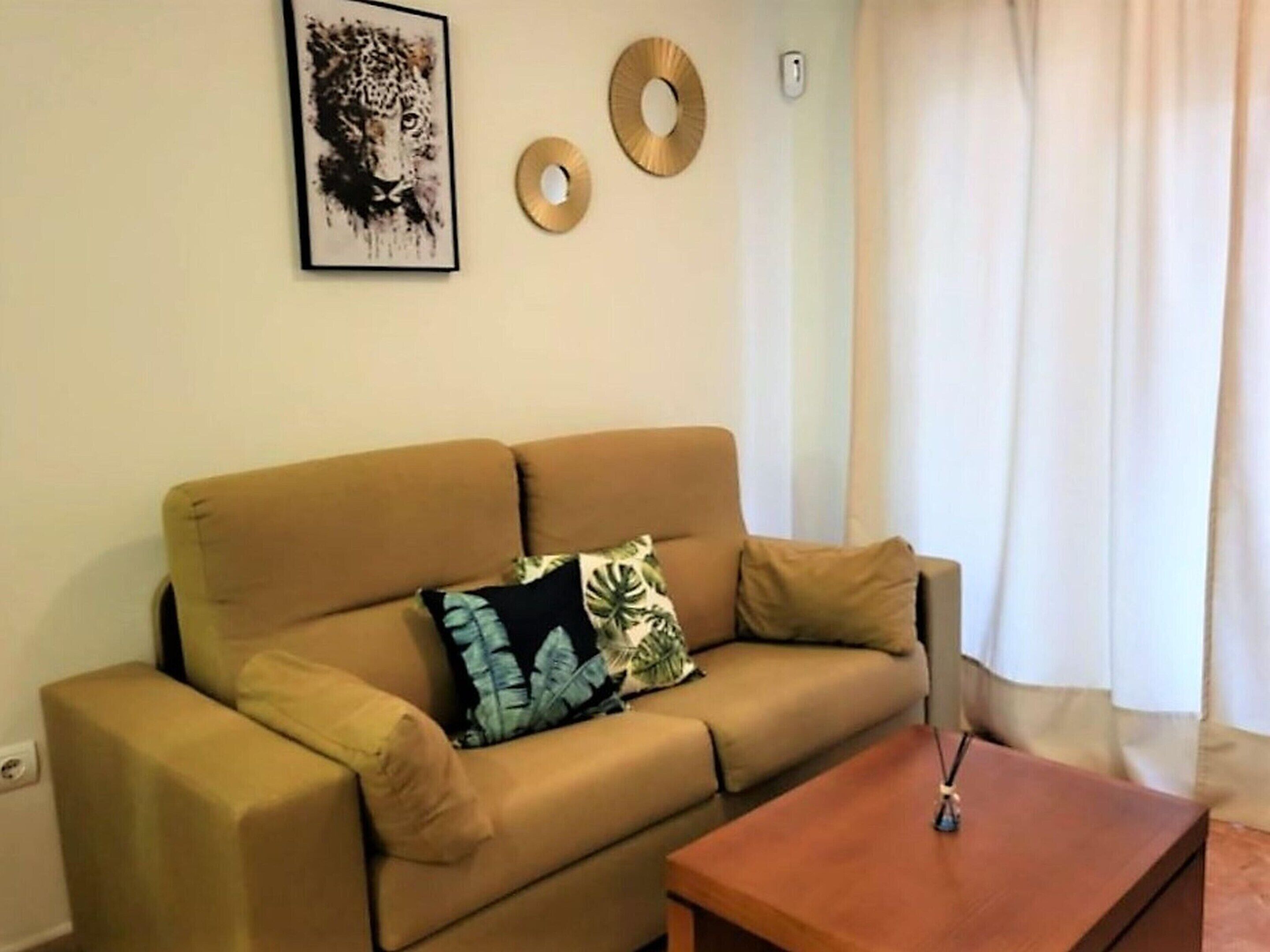 Ein Wohnzimmer mit beiger Couch, zwei Kissen, einem Couchtisch und Wanddekorationen, darunter ein gerahmtes Bild und zwei runde, goldfarbene Spiegel.
