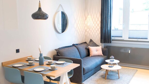 Ein modernes Wohnzimmer mit Essbereich, einem grauen Sofa, einem runden Spiegel und einem Fenster mit Vorhängen.