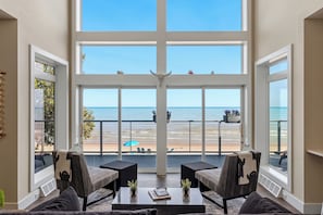 A room with a large window overlooking a beach, two armchairs, a coffee table, and a view of the ocean. - Modern | Beachfront | Sleeps 10 (Oostburg)