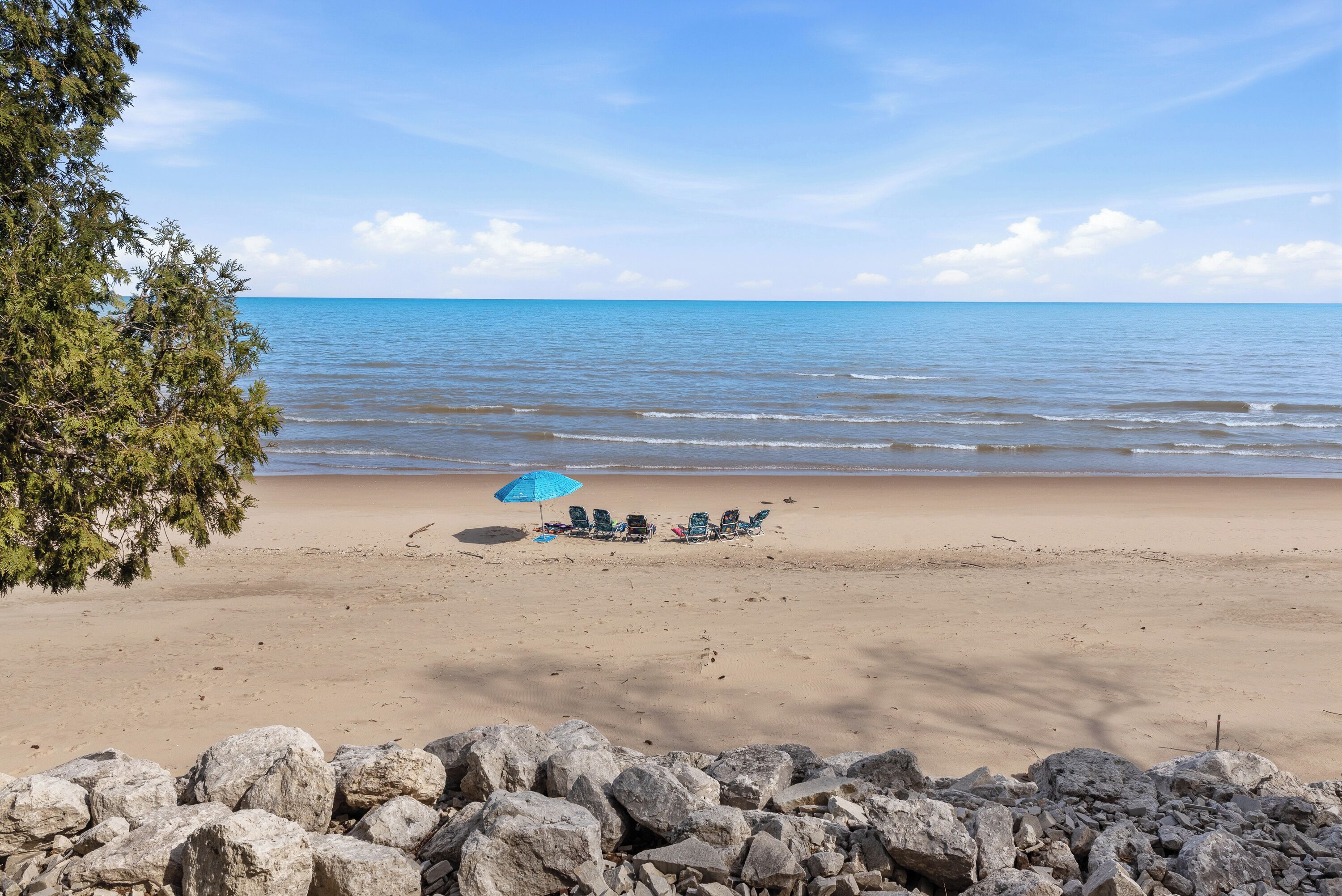 Une plage avec une rangée de chaises sous un parasol bleu, un premier plan rocheux et un ciel dégagé.