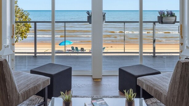 A room with a large window overlooking a beach, two armchairs, a coffee table, and a potted plant. - Modern | Beachfront | Sleeps 10 (Oostburg)