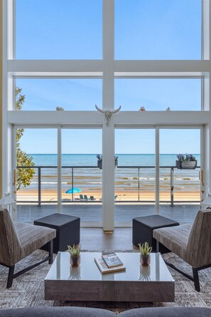 Ein Zimmer mit großem Fenster mit Blick auf einen Strand, zwei Sessel, ein Couchtisch und ein Blumentopf.