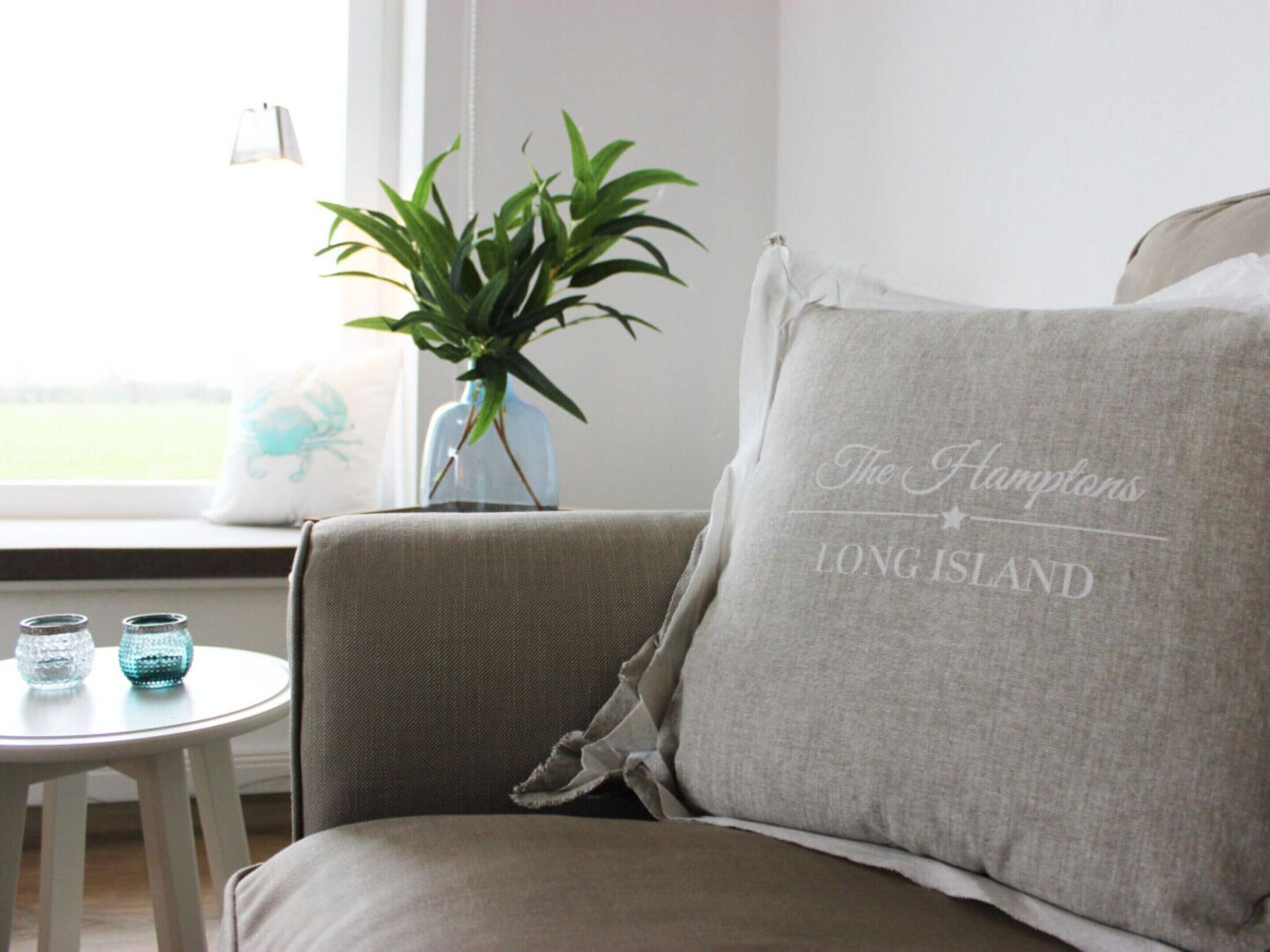 A room with a grey sofa, a small white table with decorative glass items, a potted plant, and a window with a view of a green landscape.