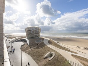 Un bâtiment moderne doté d’un design géométrique original, situé près d’une plage, avec une promenade et une structure circulaire.