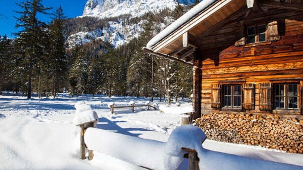 A wooden cabin with a snow-covered roof, surrounded by a snowy landscape and a mountain in the background.