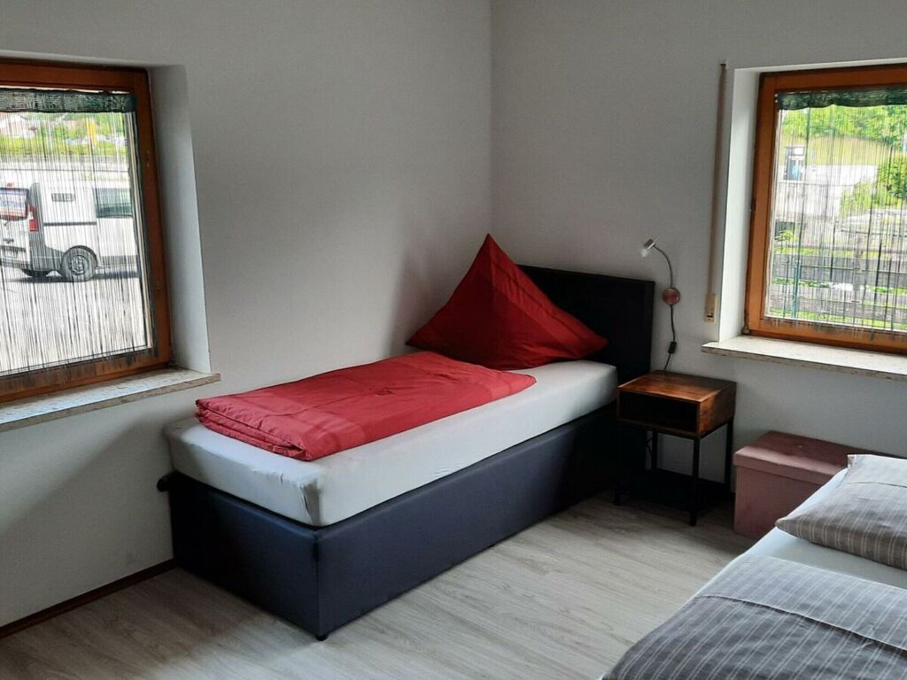 A compact bedroom with a single bed, a nightstand, a small bedside table, and two windows.