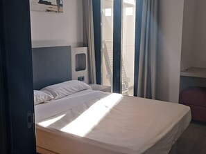 A hotel room with a bed, a small bedside table, a window with curtains, and a view of the outside.