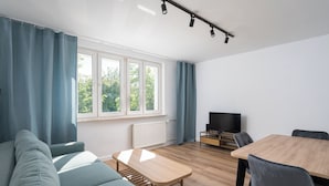 A modern living room with a sofa, a wooden coffee table, a dining table with chairs, a television on a stand, and large windows with curtains. - Ochota Comfort Apartment (Warszawa)