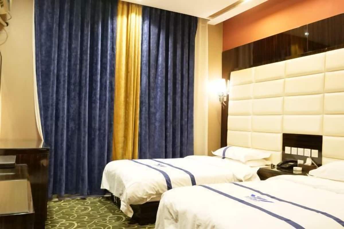 Una habitación de hotel con dos camas, un ventanal con cortinas, cabecero y una mesita de noche con lámpara.