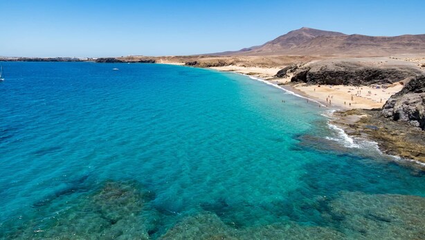 A coastal beach with clear turquoise waters, rocky shorelines, and a distant mountain. - "Oasis" Apartment in Playa Blanca | Lanzarote - Spain (Playa Blanca)