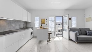 A modern kitchen with white cabinets, a central island, and a grey sofa in a bright living area with large windows. - "Oasis" Apartment in Playa Blanca | Lanzarote - Spain (Playa Blanca)