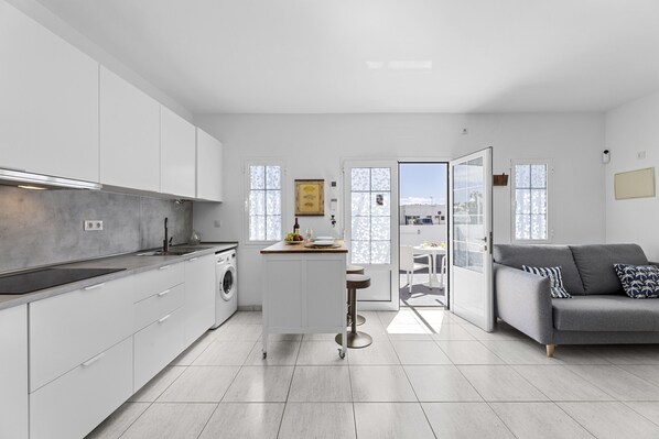 A modern kitchen with white cabinets, a central island, and a grey sofa in a bright living area with large windows.