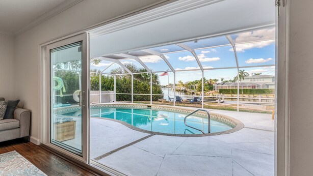 Zona de piscina con puerta de vidrio que da a un patio, piscina y vista a una casa y palmeras.