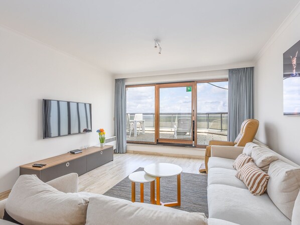 Une chambre d’hôtel moderne dotée d’un grand balcon, d’un téléviseur à écran plat, d’un canapé, d’une table basse et d’une table d’appoint agrémentée d’un vase de fleurs.