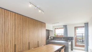 Une cuisine moderne avec un mur recouvert de panneaux en bois, un îlot central doté d’un évier et un coin repas avec une longue table et des chaises.