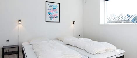 A bedroom with two beds, a small bedside table, a wall-mounted light, and a window with a view of buildings.