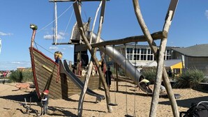 Une aire de jeux de plage avec une structure ludique sur le thème d’un navire, comprenant des toboggans et des balançoires.