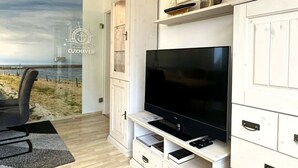 Un salon moderne doté d’un grand téléviseur à écran plat, de meubles blancs et offrant une vue sur la plage grâce à une porte vitrée.