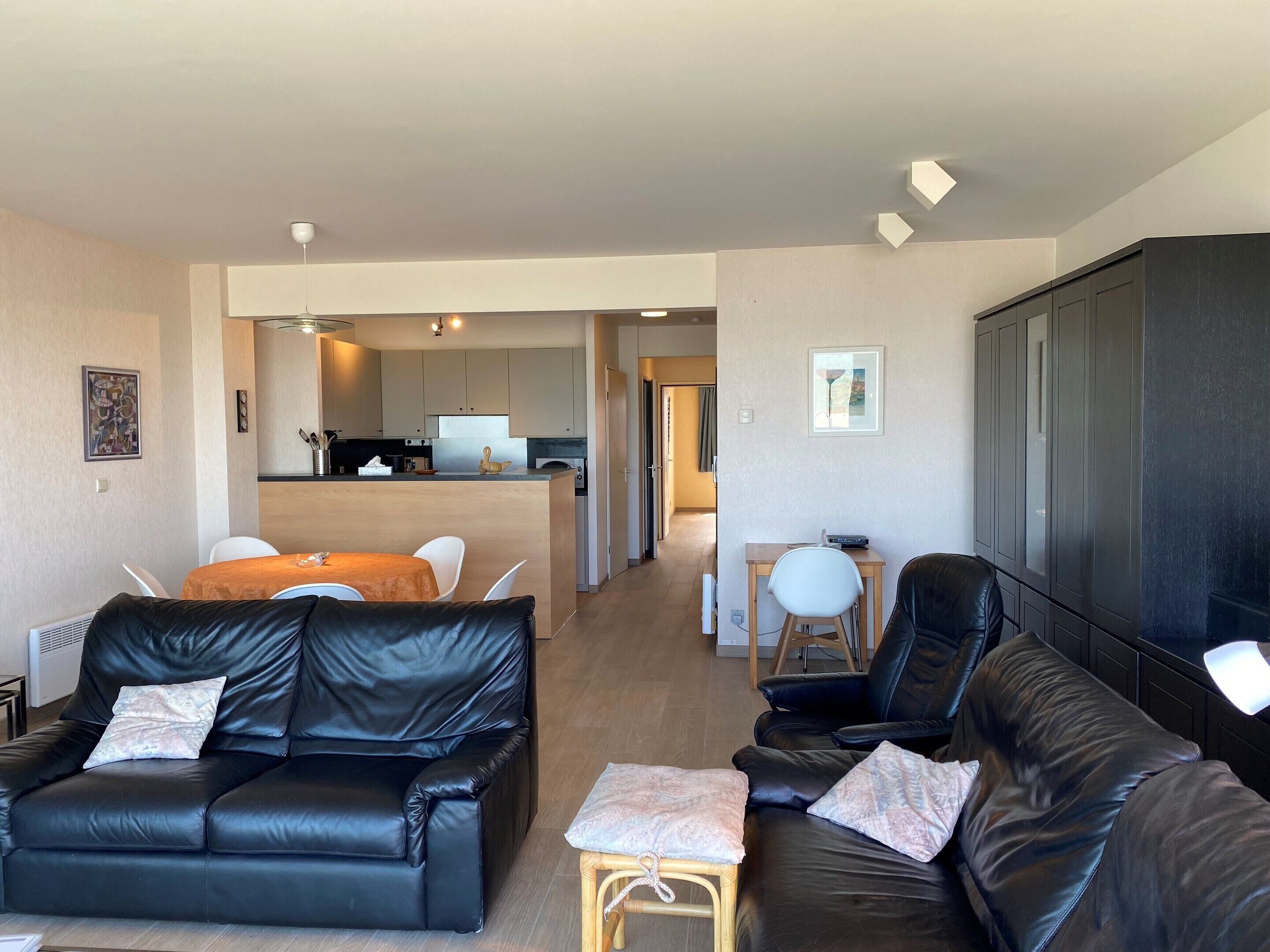 A modern living room with black leather sofas, a wooden dining table, and a kitchen in the background.