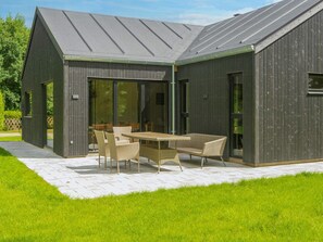 A modern building with a patio area featuring a table and chairs, large windows, and a grassy lawn. - 4 Star Holiday Home in Glesborg (Glesborg)