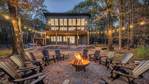 Ein modernes Haus mit Terrasse, umgeben von Adirondack-Stühlen und einem Feuerkorb, gelegen im Wald.