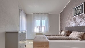 Une chambre à coucher comprenant un lit, une commode, un banc, un radiateur et une fenêtre avec des rideaux légers.