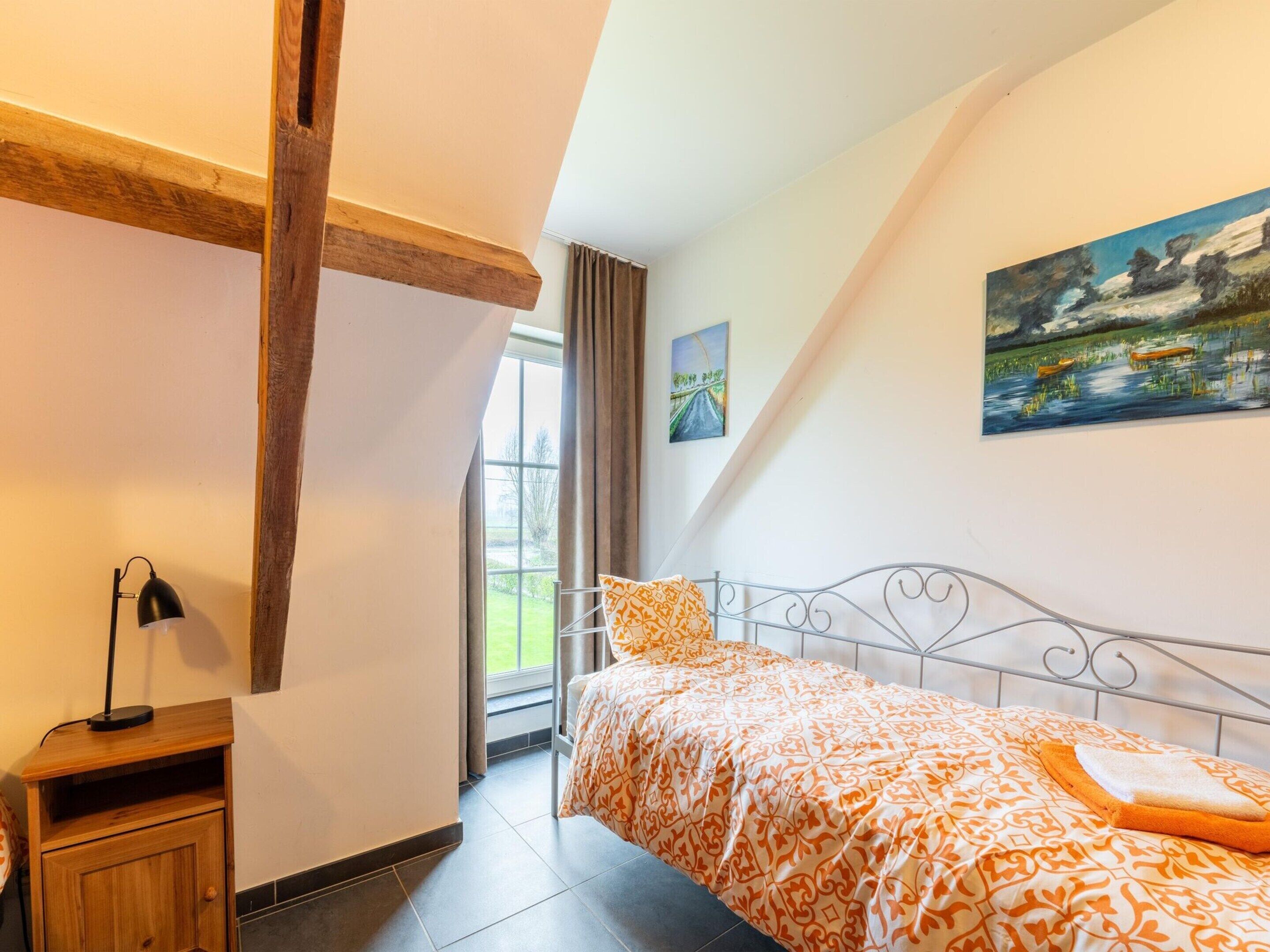 Quarto com cama, mesa de cabeceira, luminária, janela com cortinas e duas pinturas na parede.