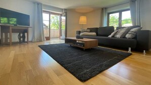 A modern living room with hardwood flooring, a black rug, a wooden coffee table, a black sofa, and a TV on a stand. - Holiday Apartment Lucky guy (Oberstaufen)