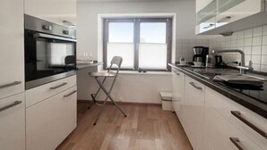 A compact kitchen with white cabinets, a foldable chair, a window with blinds, and a coffee maker. - Holiday Apartment Lucky guy (Oberstaufen)
