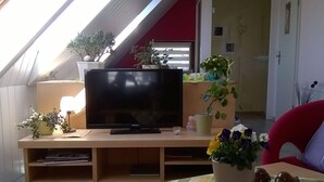 A living room with a flat-screen TV on a wooden stand, surrounded by potted plants and a checkered tablecloth. - Apartment at the Knappensee in Gross Sarchen (Ebersbach-Neugersdorf)