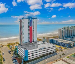 A high-rise hotel with a parking garage, beachfront view, and a clear sky.