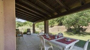 Un patio techado con una mesa dispuesta para cuatro personas, rodeado de sillas blancas, con vistas a un área verde con árboles.