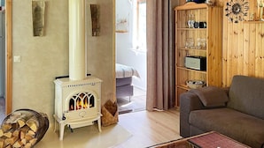 Una sala de estar acogedora con una estufa de leña blanca, un sofá marrón, una mesa de centro de cristal y una estantería de madera con diversos objetos.