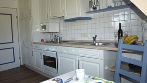 A kitchen with white cabinets, a microwave, a stove, and a sink. There is a blue shelf above the sink with kitchen utensils. - Kille Family - Attic (St. Peter-Ording)