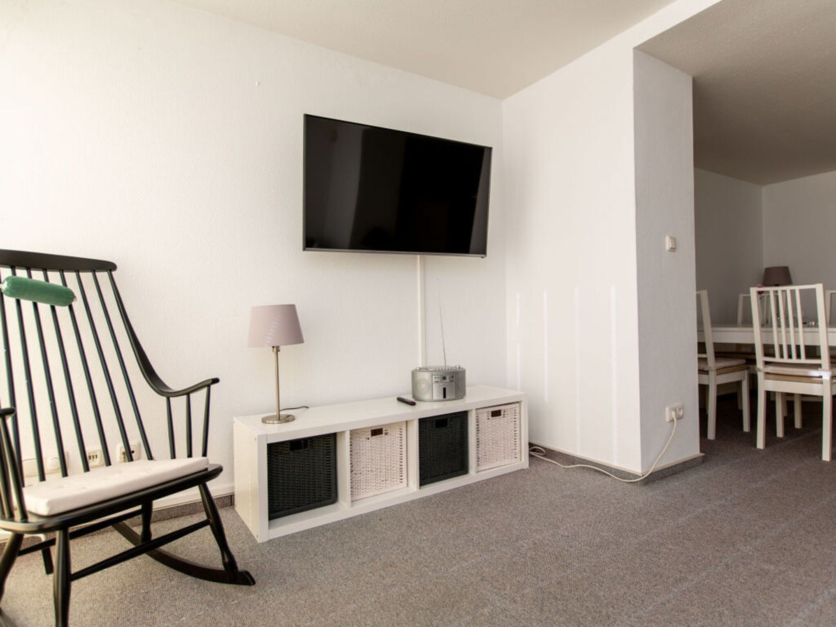 A room with a television mounted on the wall, a white console table with a lamp, a rocking chair, and a dining area with chairs.