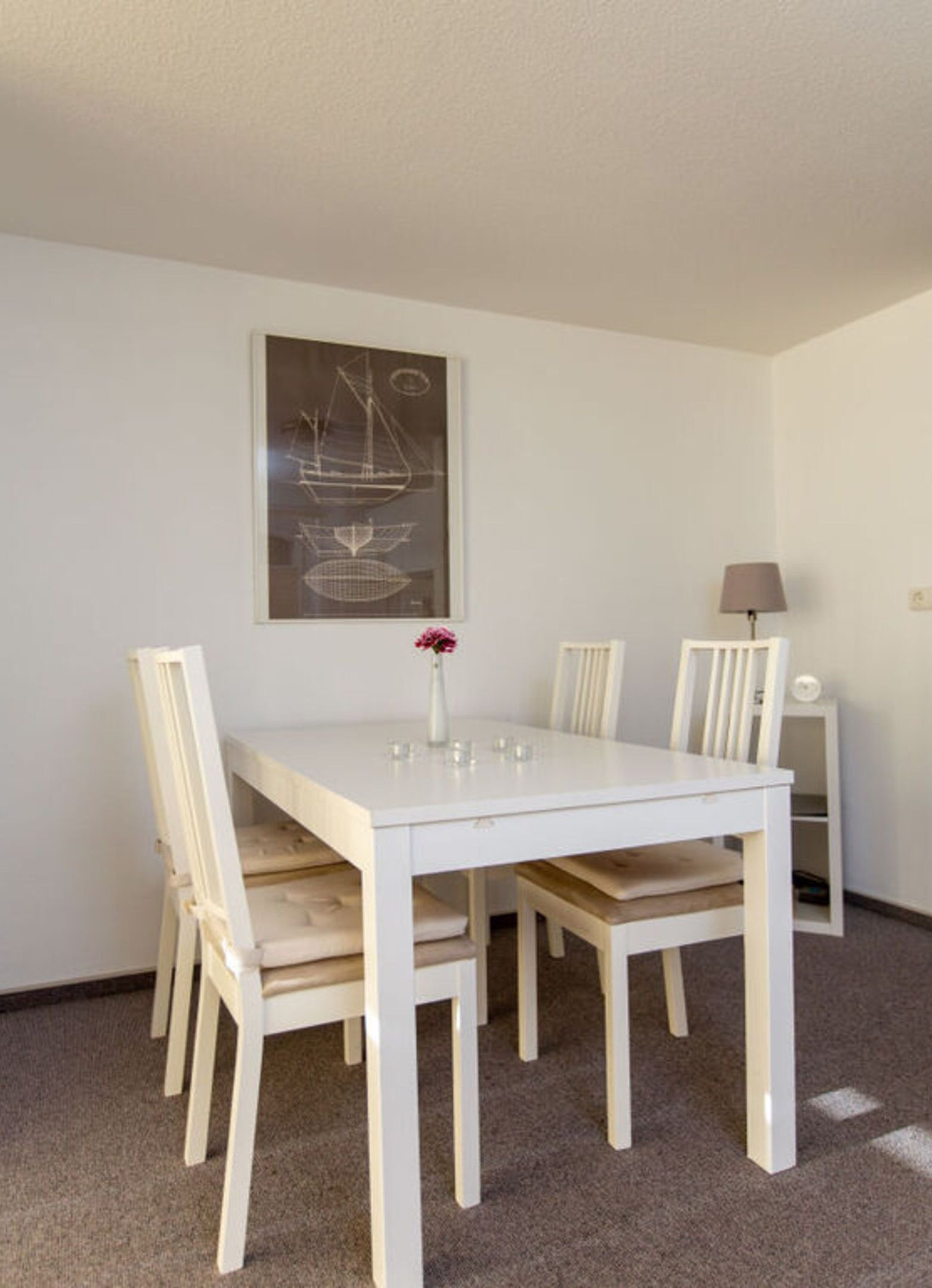 A dining area with a white table and chairs, a vase with flowers, and a framed picture on the wall.