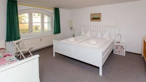 Une chambre avec un lit blanc, une fenêtre avec des rideaux verts, un radiateur, une petite table de chevet avec une lampe et un tableau au mur.