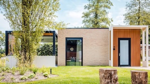 Een modern gebouw met een bakstenen gevel, een glazen deur en een houten deur. Er zijn bomen en een gazon met houten boomstronken.