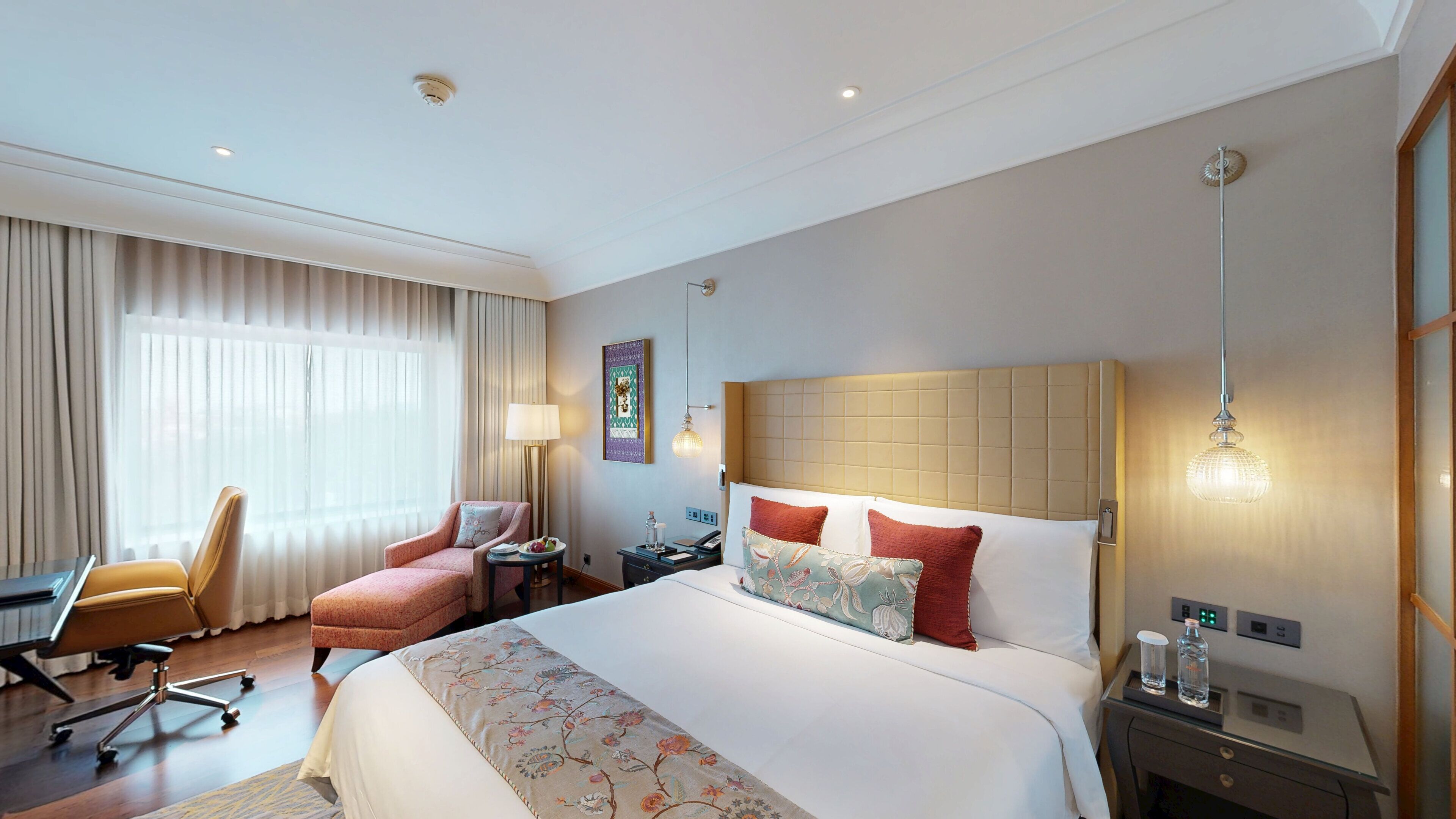 A hotel room with a large bed, a desk, a chair, a lamp, and a bedside table.