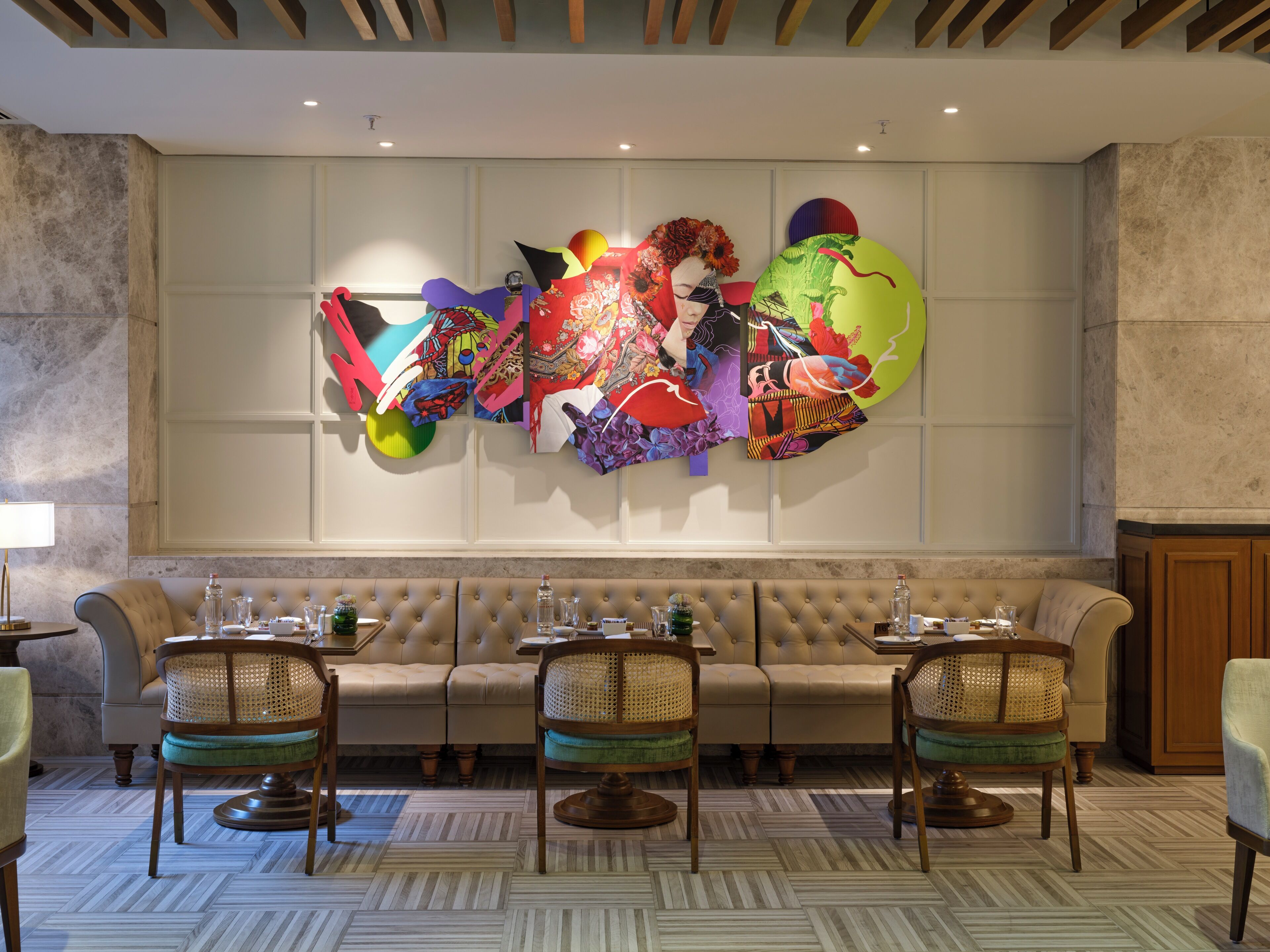 A modern hotel lobby with a large, colorful abstract art piece on the wall, a plush beige sofa, and three wicker chairs with round wooden bases.