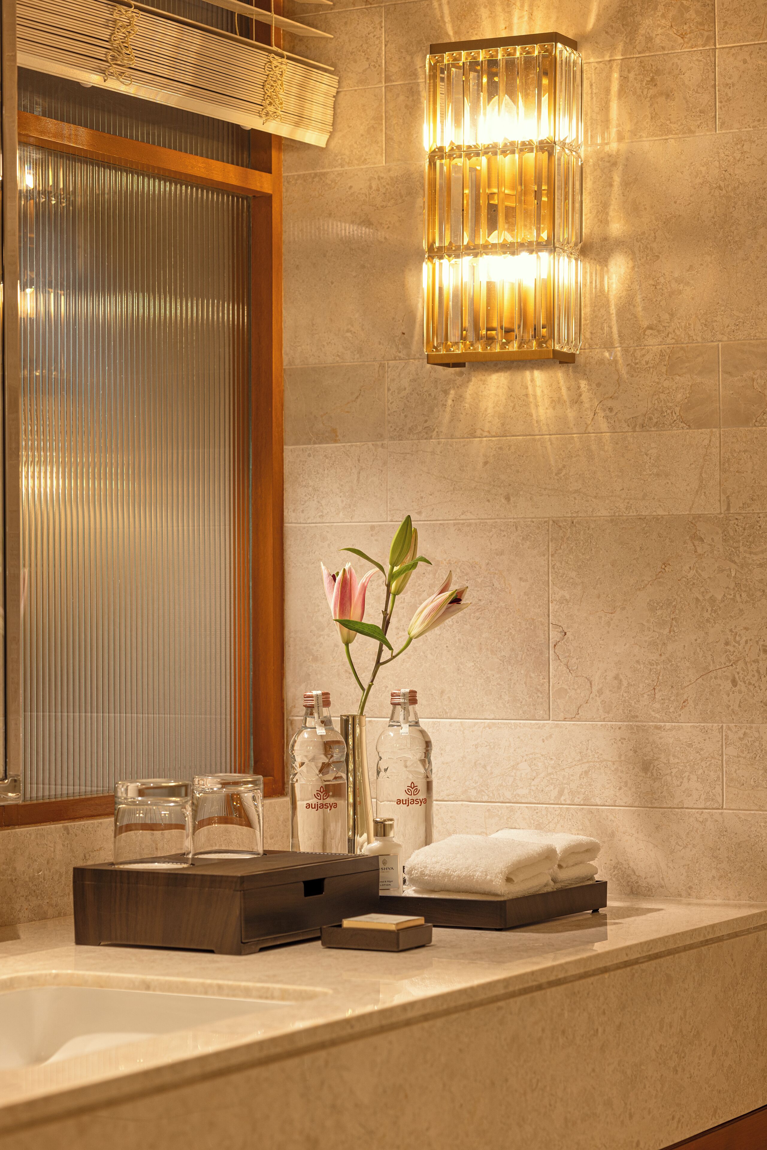 Un baño moderno con un espejo grande, una lámpara fijada a la pared, un jarrón con una flor rosa y artículos de tocador ordenados.