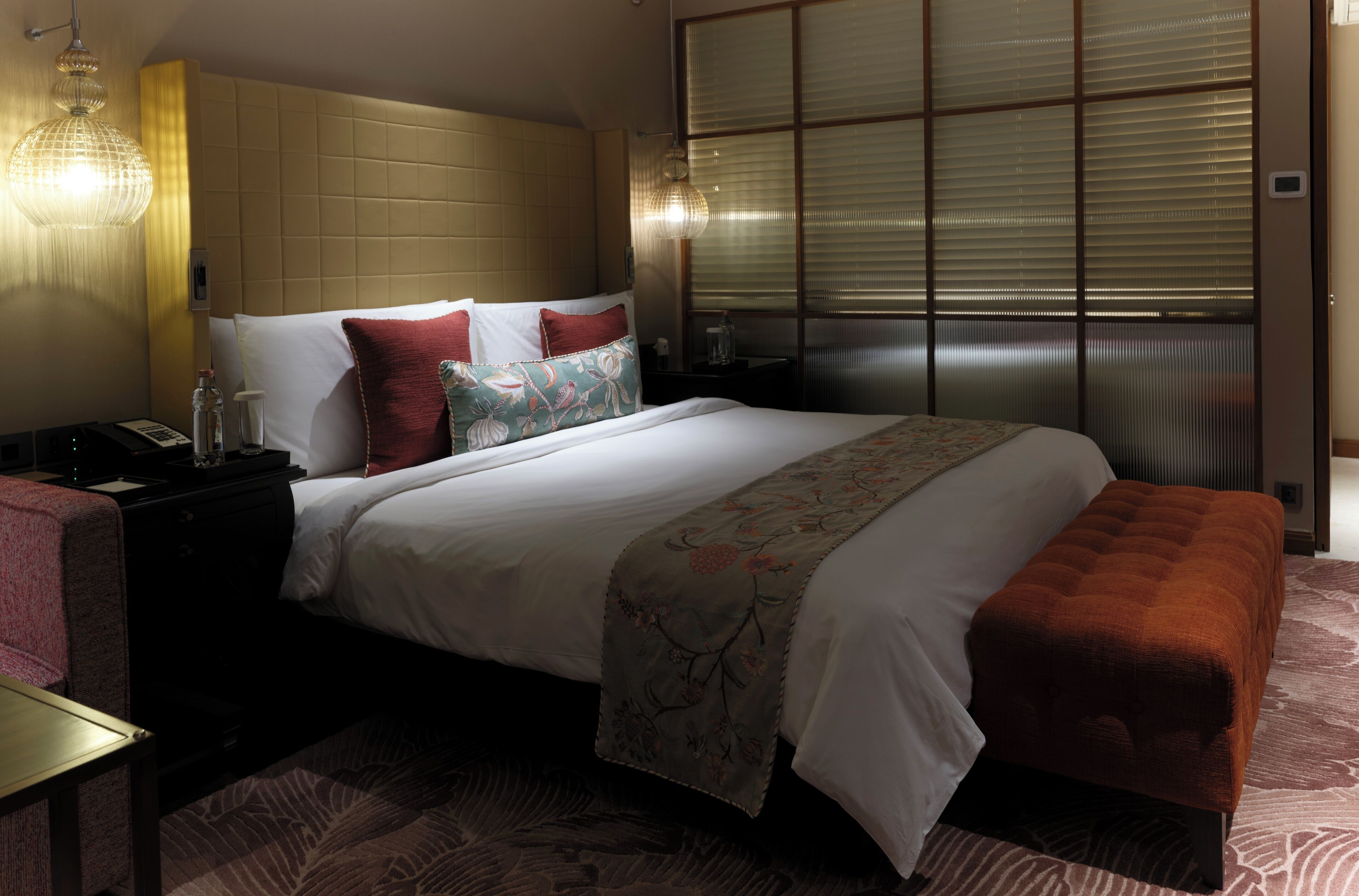 A hotel room with a large bed, a bedside table with a lamp, a nightstand, and a window with blinds.