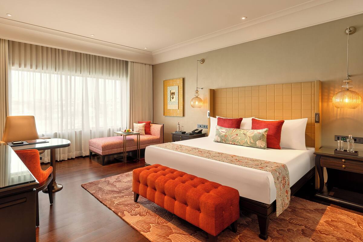 Habitación de hotel con una cama grande, una zona de estar con una mesa y sillas, un escritorio con lámpara y un ventanal con cortinas.