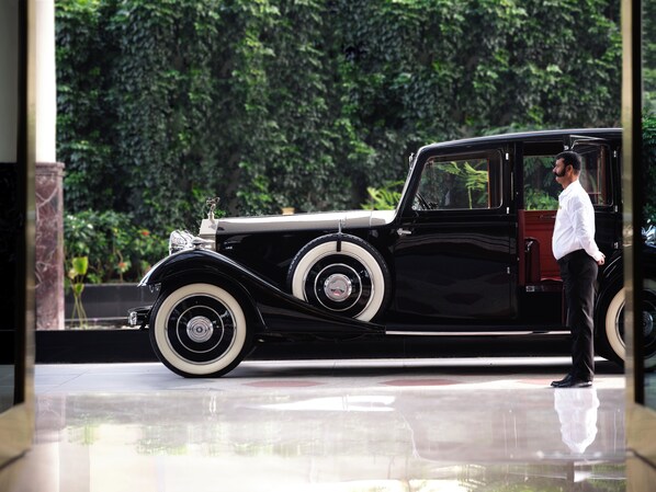 Un coche clásico negro con ruedas blancas estacionado en un área bien iluminada, con piso de mármol y follaje al fondo.
