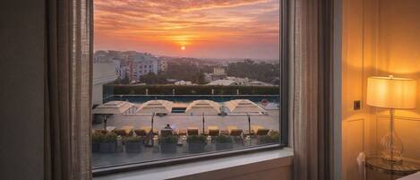 A hotel room with a view of a sunset over a cityscape, a balcony with sun loungers, and a lamp on the bedside table.
