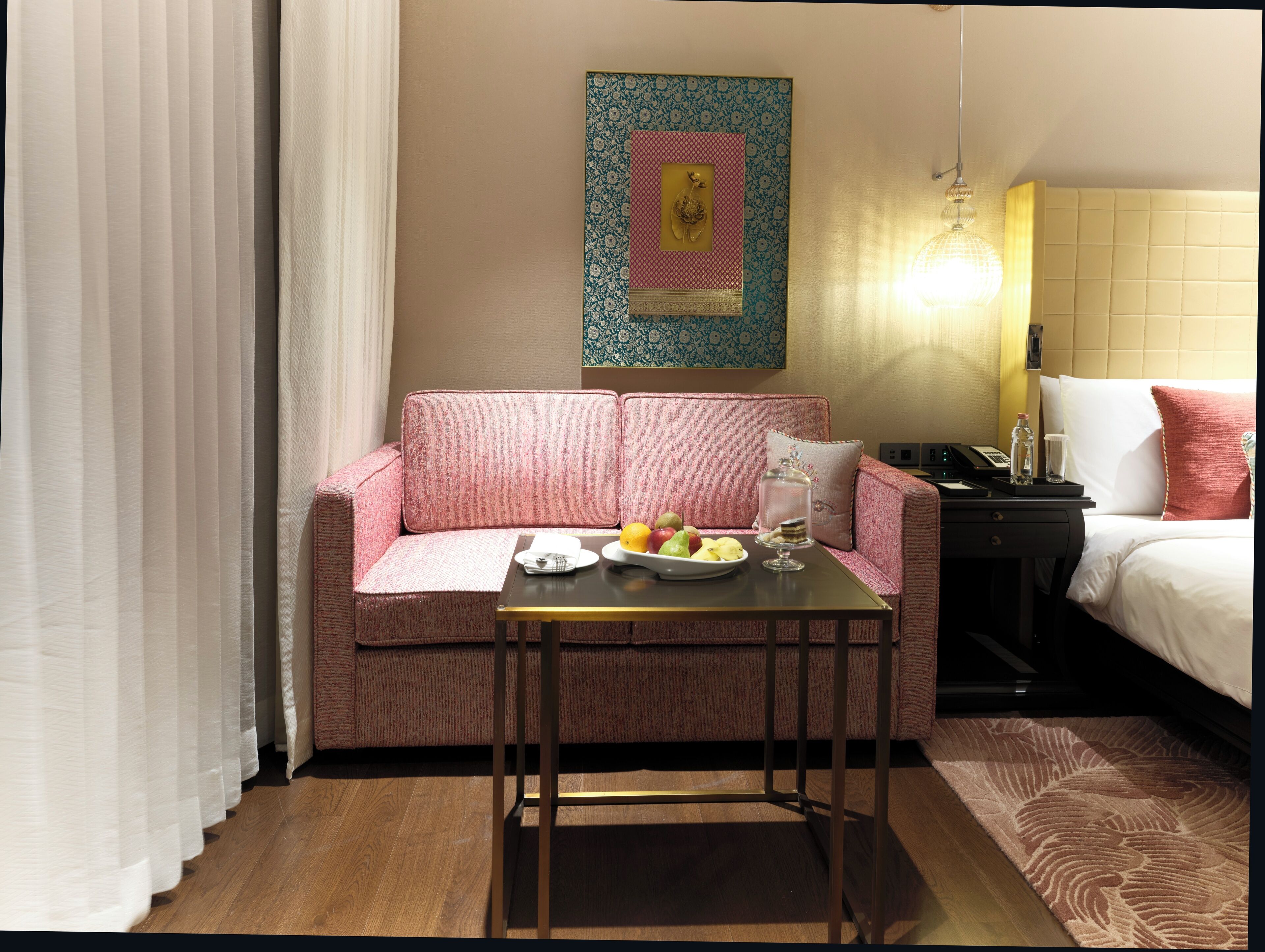 A hotel room with a pink sofa, a small table with fruits and a glass, a bed with white bedding, and a decorative wall piece.